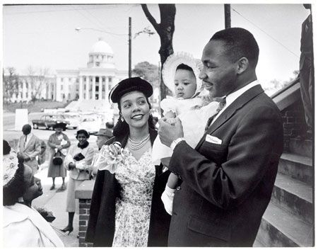 Dr. Martin Luther King Jr. holds his infant daughter as he stands beside his wife smiling.